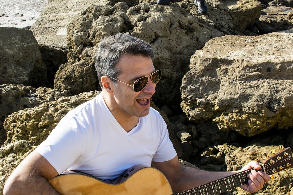 Guitariste de ROCK'ELLE souriant en jouant de la guitare acoustique sur un fond de rochers côtiers à La Rochelle