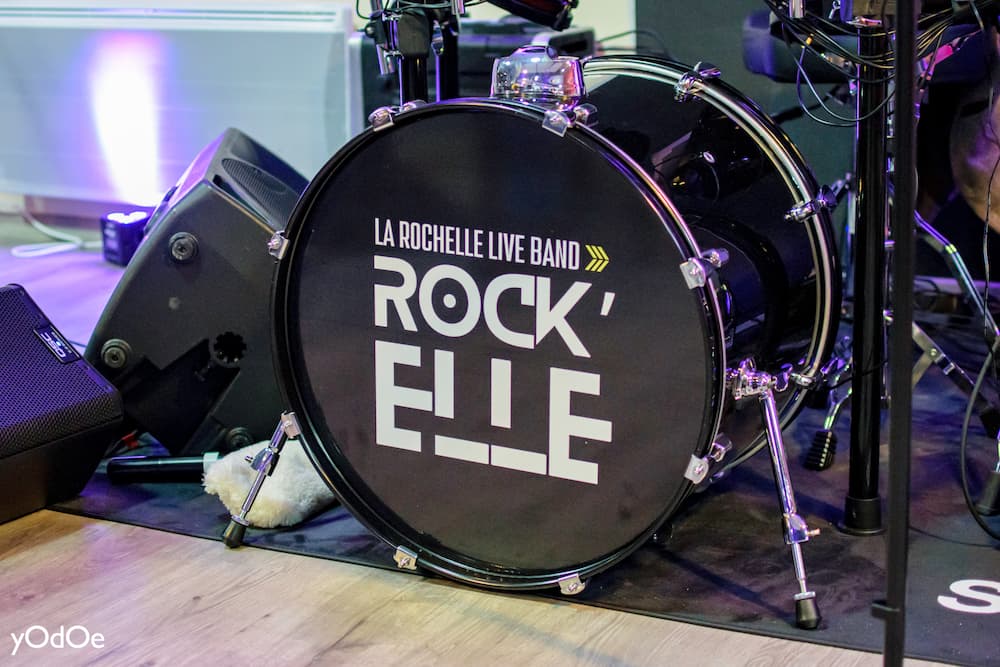 Chanteuse de ROCK'ELLE captivant l'audience avec sa performance, micro en main, avec un musicien flou en arrière-plan.