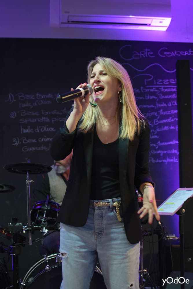 La chanteuse de ROCK'ELLE s'anime devant le micro avec les membres du groupe détendus sur un canapé en plein air.