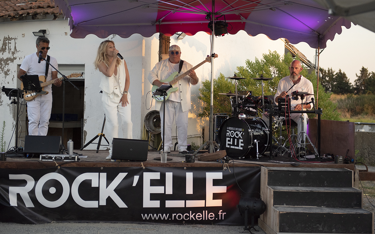 Guitariste de ROCK'ELLE souriant en jouant de la guitare acoustique sur un fond de rochers côtiers à La Rochelle