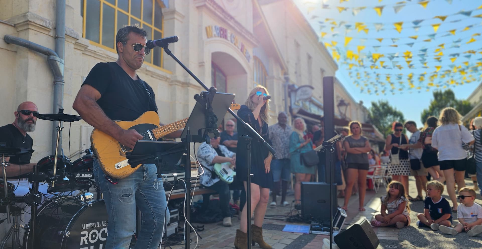 Guitariste concentré de ROCK'ELLE jouant sur scène avec un batteur en arrière-plan pendant un concert en direct.