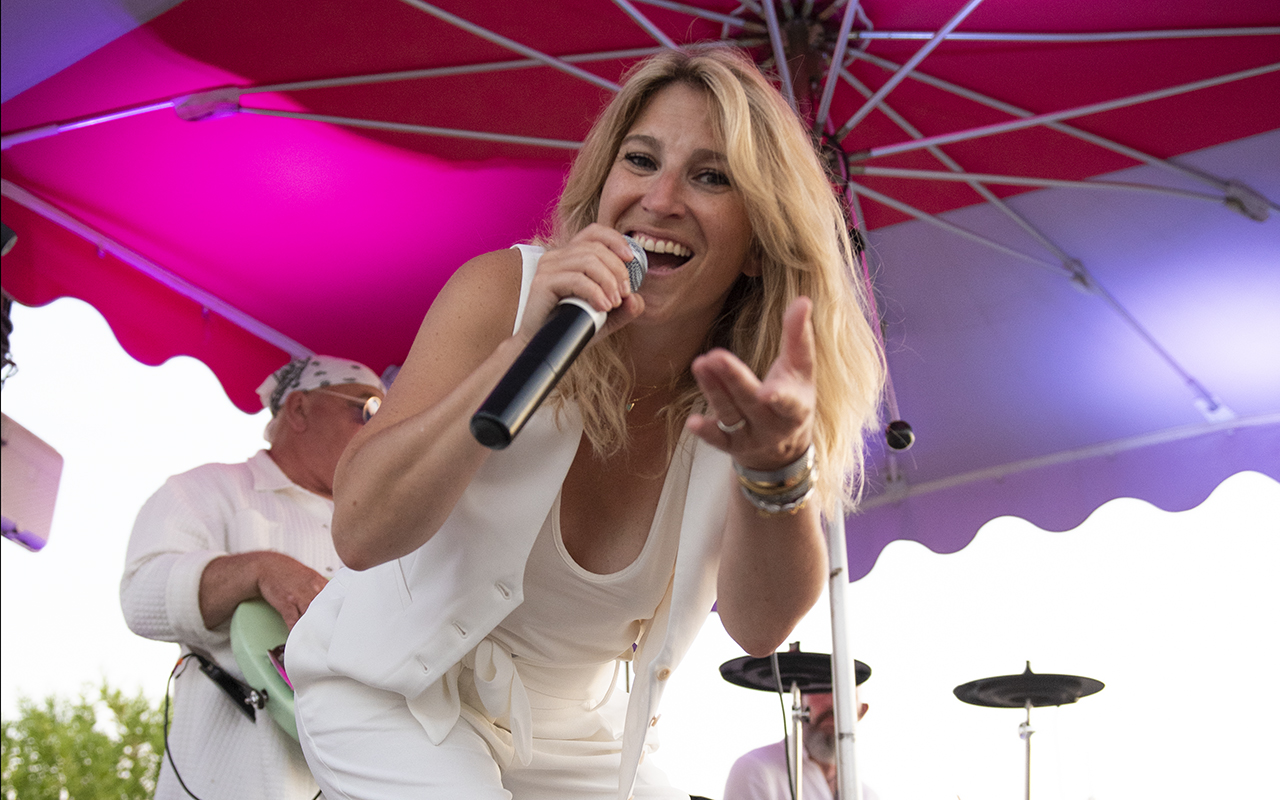 Chanteuse de ROCK'ELLE captivant l'audience avec sa performance, micro en main, avec un musicien flou en arrière-plan.